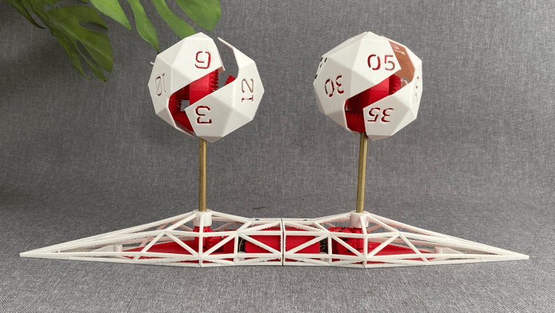 An angular, 3D-printed base holds two icosahedra with numerals on their faces. Each icosahedron has a zig-zagging path running through it, showing red gears inside.