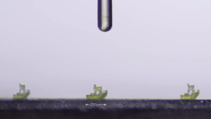 A white background is shown, with a grey metal plate at the base of the image. On the plate are three tiny green Benchy models. Above the Benchies is a glass cylinder. Below one of the Benchy models, text says "250 µm".