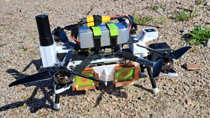 A drone is shown, carrying underneath it a white plastic box. On the side of the box are two patch antennas. A camera extends from one end of the box, and a large GPS antenna from the other end.