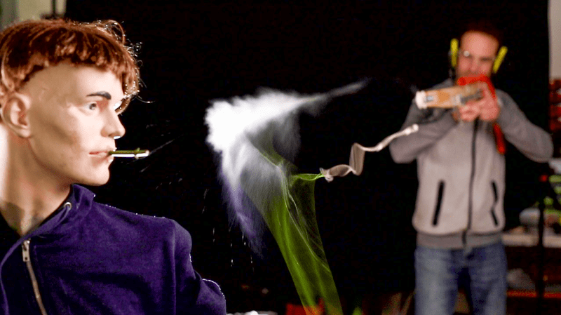 On the left side of the image, a mannequin holds a blade of grass in its mouth. A trail of dust follows a blurred green trail past the piece of grass. A man in the background is pointing a wooden device toward the mannequin.