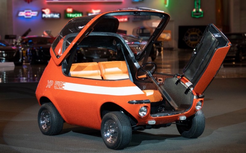 A small orange car with a bold horizontal white stripe and the text "XP 512E" emblazoned on the side. The footprint of the vehicle barely exceeds the size of the seats, almost like an Isetta, but the top canopy opens upward and the lower front fascia contains a side opening door. If it were green, it would probably be what an LLM would generate with the prompt, "Make a car that looks like a frog."