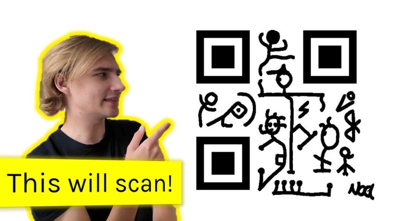 A person with blonde hair and light skin in a black t-shirt points at an image of a large QR code with what appear to be pictographic humans dancing in place of the usual blocky squares between the finding squares.