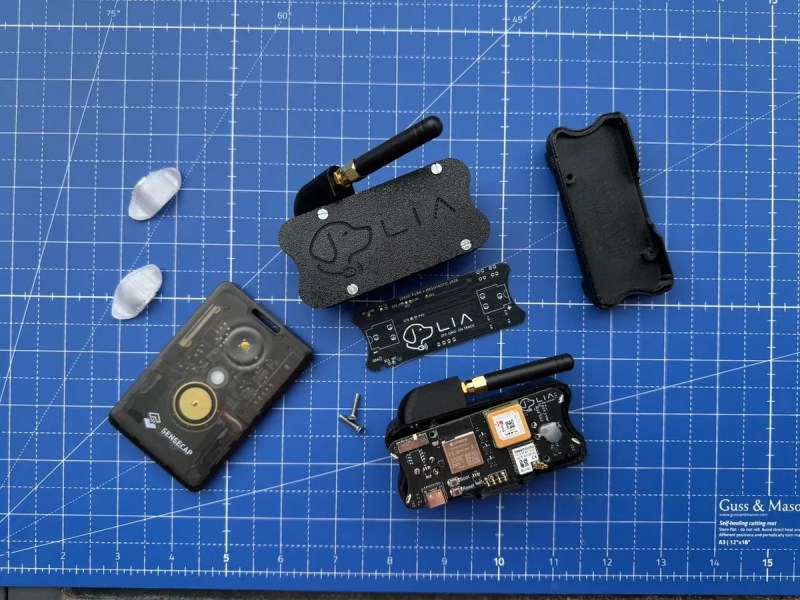 Four black plastic devices, two with a black, stubby antenna, sit on a blue cutting mat with white guidelines. There is also a PCB and a couple grey-ish clips in the assortment. One of the devices is open, showing another PCB with various components including a GPS antenna and LoRa radio. A silhouette of a dog is screen printed next to the text "LIA" on the PCBs, and the same text and image is present in the shell of the device in the top center.