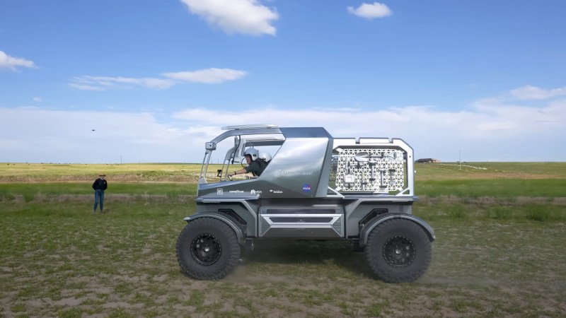 A shiny, silver vehicle with tools on a mesh rack on the back half and a large open cab up front drives across a grassy field under blue, partly cloudy skies. A person in a black shirt stands in the background with a drone hovering above their head. Two women in white helmets are piloting the vehicle from the cab.