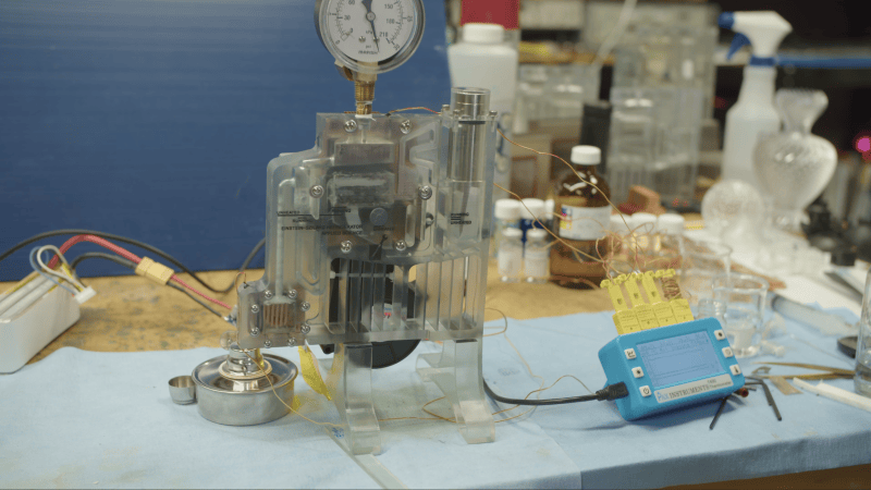 A clear plastic block is shown on a workbench. The plastic block has channels running through it, with liquid flowing through the channels. A pressure gauge is mounted to the top of the block, and an alcohol burner is placed near the base.