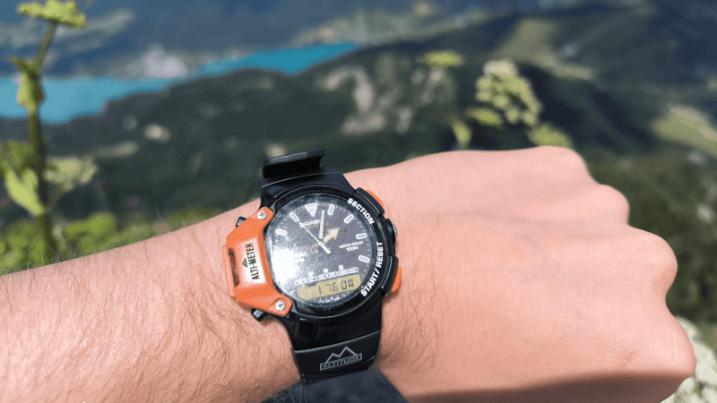 A man's arm is shown against a mountainous backdrop. The man is wearing a black watch with an orange segment on one side.
