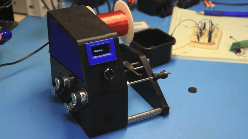 A black 3D printed machine sits on a workbench. A coil of copper wire is suspended from one side of it, and a screen on the front of it reads "Homing".