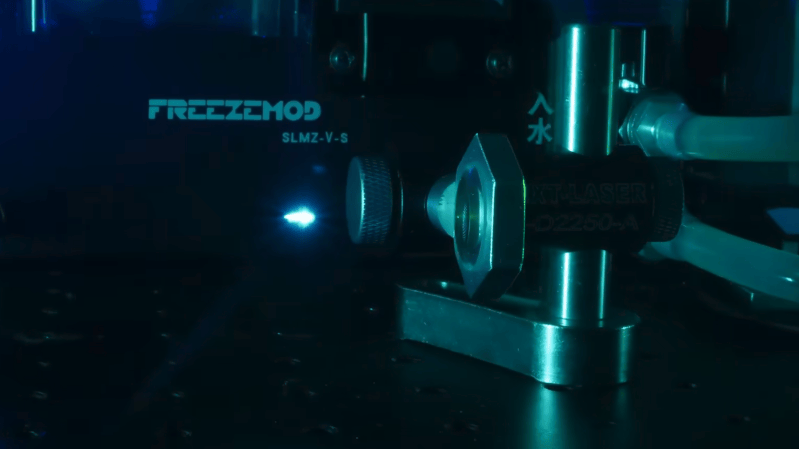 A dark optical bench is shown, with a lens in the right-hand part of the image. In front of the image, a bright blue spark appears in the air, with no equipment near it.