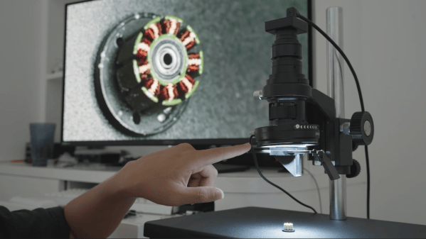 A man's hand is seen adjusting a lever connected to a lens, which is pointing down towards the windings of a small motor. A screen behind this shows an image of the windings, taken from an angle slightly to the side.