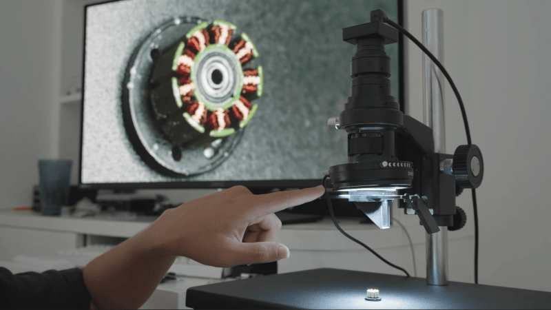 A man's hand is seen adjusting a lever connected to a lens, which is pointing down towards the windings of a small motor. A screen behind this shows an image of the windings, taken from an angle slightly to the side.