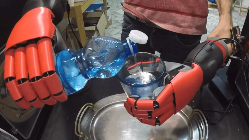 A pair of red, articulated, 3D printed hands are being used to pour water from a plastic bottle into a glass. One hand holds the bottle, and one holds the glass.